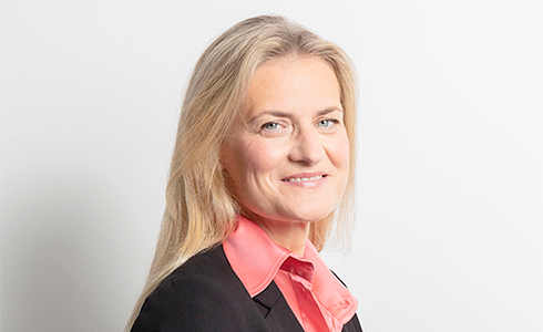 Professional headshot of a smiling blonde woman in a pink blouse and black blazer against a light background, suitable for a corporate bio or About Us page.