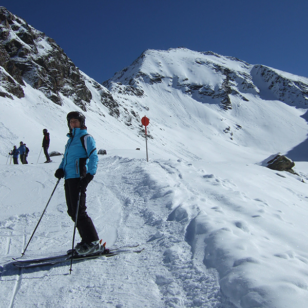 Skier in a blue jacket and helmet standing on a groomed snowy trail with jagged snow covered mountains in the background
