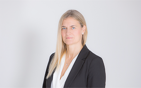 Professional woman in a black blazer and white blouse, standing against a neutral light background, looking at the camera.