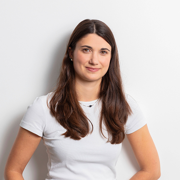 Portrait of a woman with long brown hair wearing a white t-shirt, smiling softly against a light backdrop.