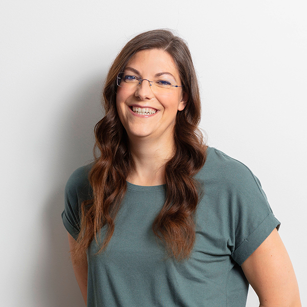 Smiling woman with long wavy brown hair and glasses, wearing a teal shirt, standing against a light background.
