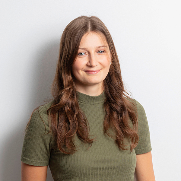 Smiling young woman with long wavy brown hair wearing an olive-green ribbed top against a light gray background.