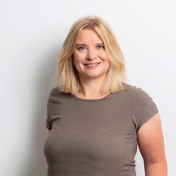 Woman with blonde hair and a taupe t-shirt smiling at the camera, against a light background.