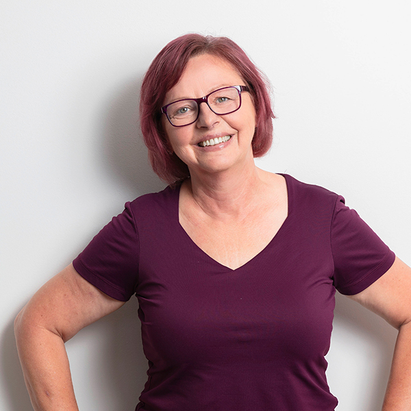 Smiling woman with short purple hair and glasses stands with hands on hips against a light wall, wearing a burgundy V-neck shirt.