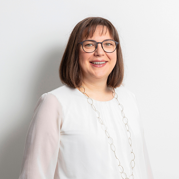 Smiling woman with short brown hair and glasses wearing a white blouse and a long circular-link necklace against a light background.