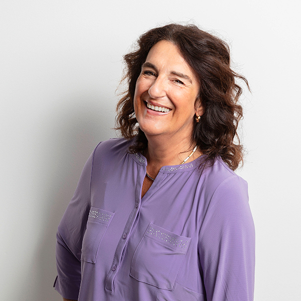 Smiling woman in a lavender blouse standing against a light background.