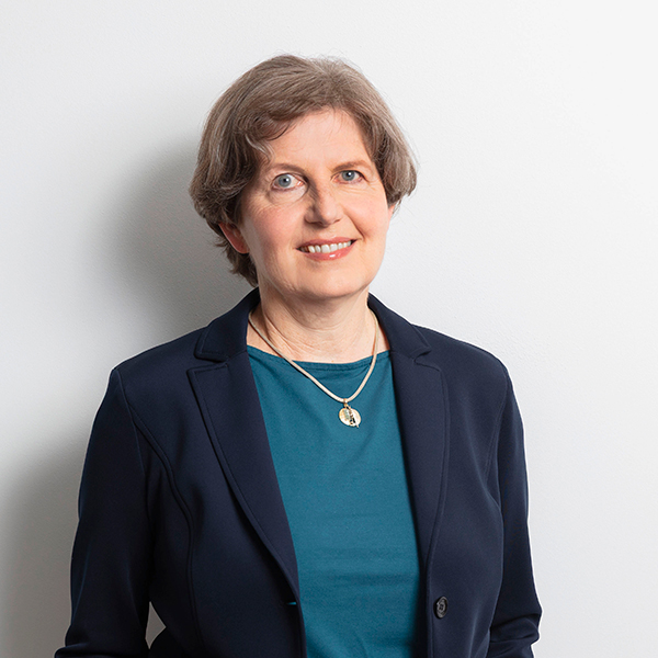 Professional head-and-shoulders portrait of a middle-aged woman with short gray hair, wearing a navy blazer over a teal blouse and a pendant necklace, smiling at the camera.