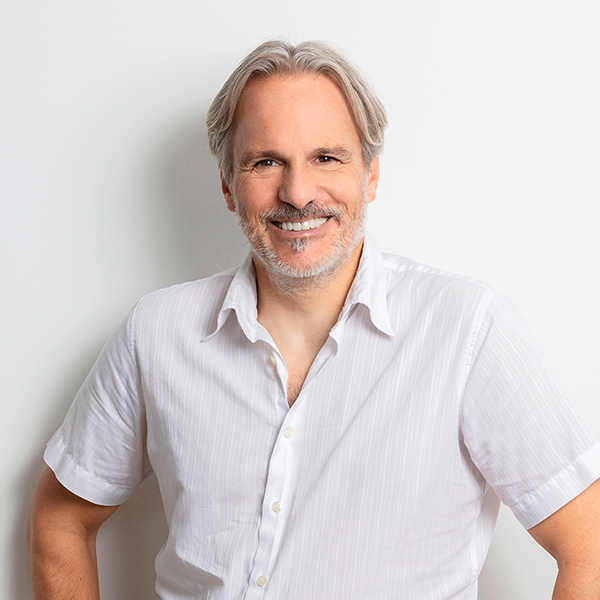 Smiling middle-aged man with gray hair and a light beard, wearing a white button-down shirt, posing against a light background.