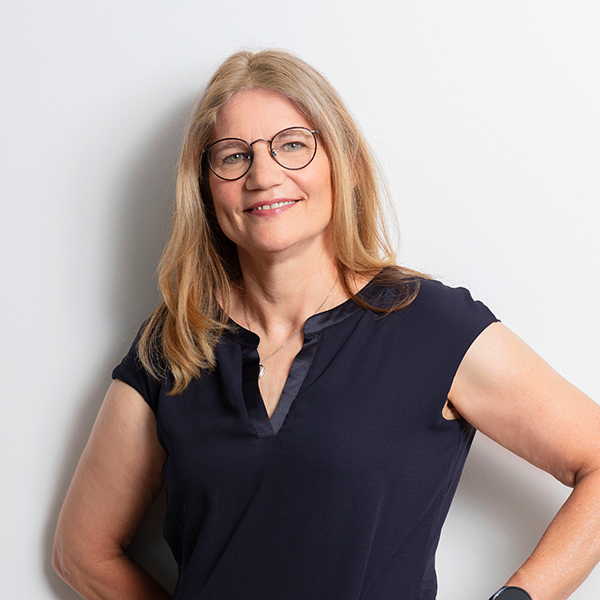 Smiling woman with round glasses in a navy blouse, standing against a white wall.