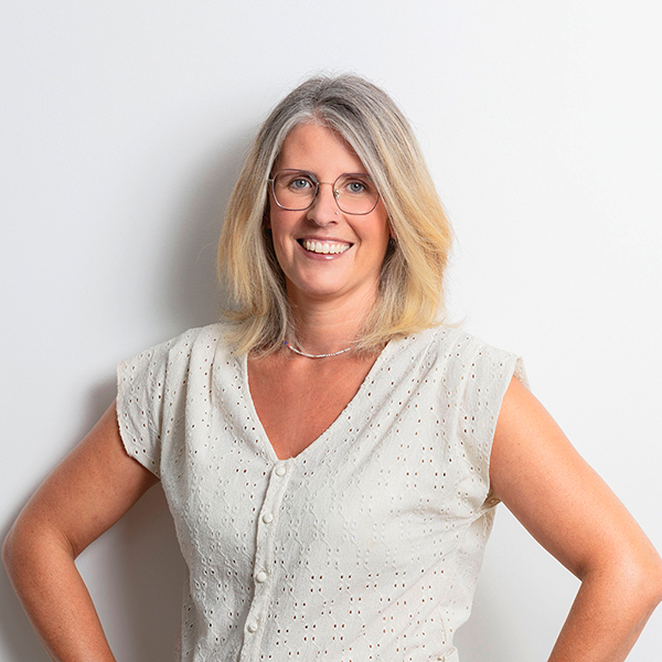 Smiling woman with shoulder-length gray-blonde hair and glasses, wearing a light eyelet blouse, posing with hands on hips against a white background in a professional portrait.