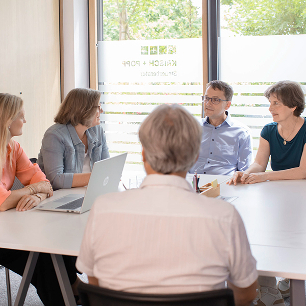 Group of coworkers in a meeting around a white conference table with a laptop and papers in a bright office with large windows