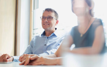 Man with glasses in a light blue shirt smiles during a meeting at a bright conference table with colleagues nearby.