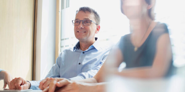 Man with glasses in a light blue shirt smiles during a meeting at a bright conference table with colleagues nearby.