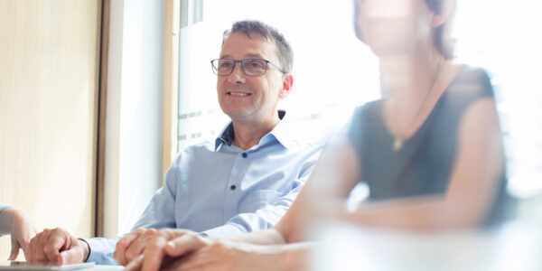 Smiling man in glasses sits at a conference table with colleagues in a bright office setting.