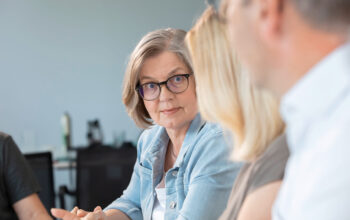 Older woman with short gray hair and glasses listens attentively to a colleague during a meeting in a bright office setting.