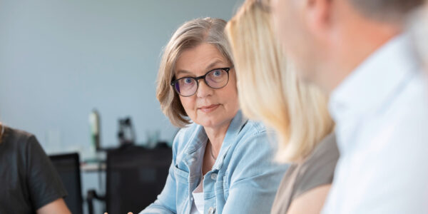 Older woman with short gray hair and glasses listens attentively to a colleague during a meeting in a bright office setting.
