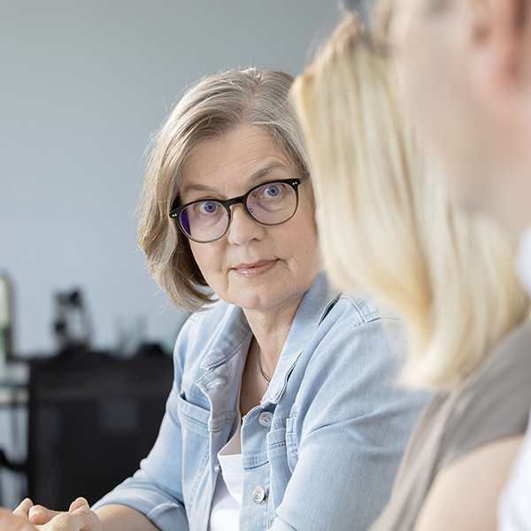 Woman with gray hair and glasses listens attentively during a meeting with two colleagues nearby in a light office setting
