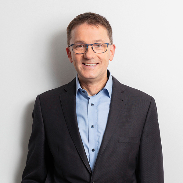 Smiling middle-aged man in a dark blazer and light blue shirt, against a light wall (professional portrait).