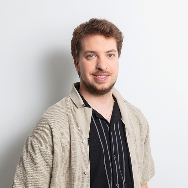 Smiling man with light brown hair wearing a black striped shirt and beige cardigan, posing against a light gray wall.