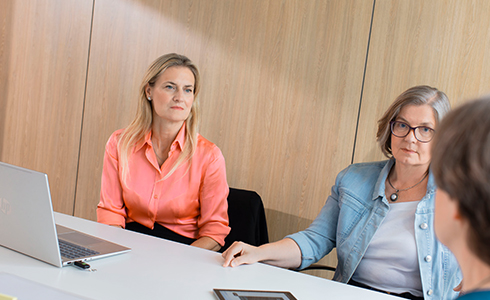 Two women sit at a conference table during a meeting; one wears a coral blouse with a laptop, the other in a light denim jacket with glasses, facing a colleague