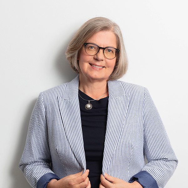 Portrait of a smiling professional woman with short gray hair and glasses, wearing a blue striped blazer over a dark top.
