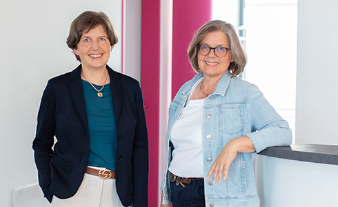Two women stand in a bright office, smiling at the camera; one wears a navy blazer with teal top, the other a light denim jacket.