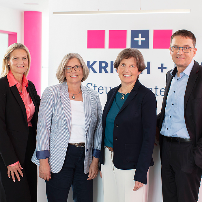 Four professionals pose for a photo in a modern office with a company sign featuring pink squares and a blue plus symbol in the background