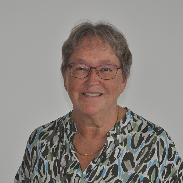 Smiling older woman with short gray hair and red-framed glasses, wearing a blue-green patterned blouse.