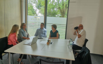 Four colleagues sit around a white conference table while a photographer on the right snaps a photo in a modern meeting room with large windows and greenery outside.