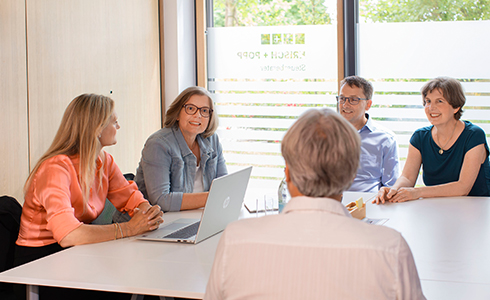 Five professionals seated around a white conference table in a bright room, discussing with an open laptop in view.