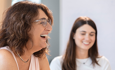 Two women smiling and chatting in a bright room, one foreground with glasses and curly hair enjoying a laugh.