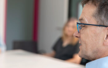 Close-up profile of a man wearing blue-framed glasses in a blurred office or meeting setting.